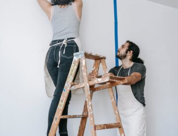 woman putting tape on wall while the man is holding the stepladder